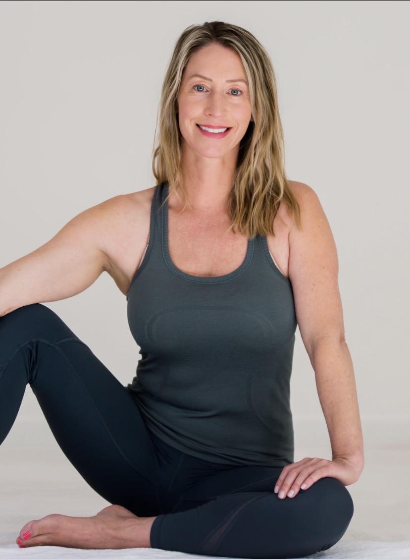 A woman with shoulder-length blonde hair seated on the floor in dark athletic wear, smiling warmly at the camera